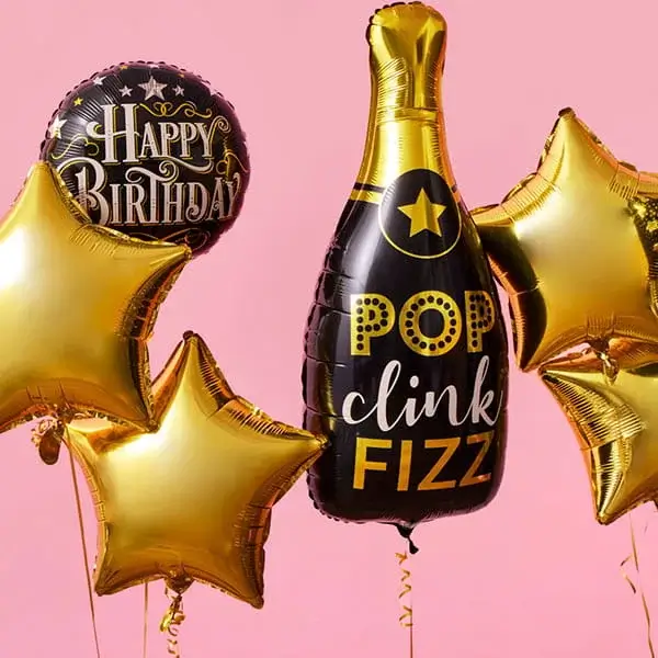 A collection of metallic balloons against a pink background, including two gold star-shaped balloons, a black round balloon with gold lettering that reads Happy Birthday, and a black champagne bottle-shaped balloon with gold accents and the words Pop Clink Fizz.